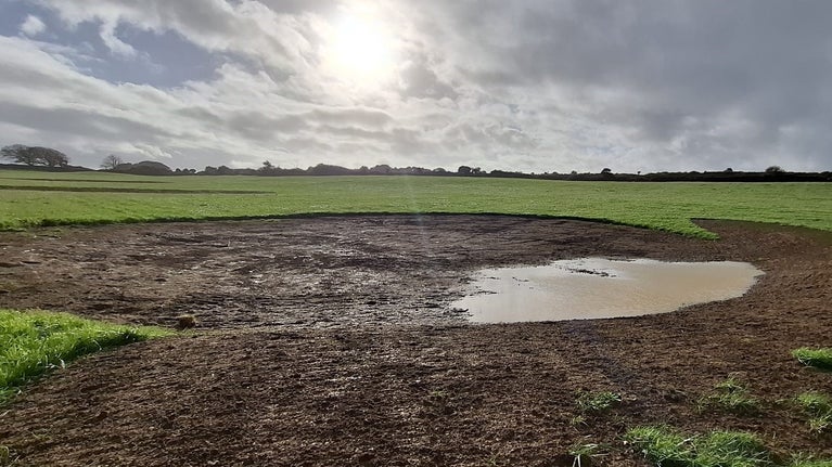 An area of a field has been excavated to form a temporary pond. It is beginning to build up water on one side. It is surrounded by fields.